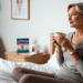 Vrouw van middelbare leeftijd kijkt nadenkend uit het raam met een kop thee, terwijl een strip met de anticonceptiepil op het nachtkastje ligt – symbool voor de overgang en hormonale veranderingen.